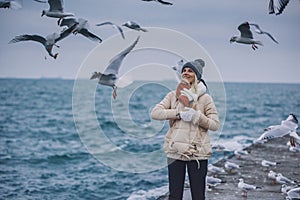 Happy young woman tourist feeds seagulls on the sea. Pretty female wearing coat, scarf, hat smiling and watching flying seagulls