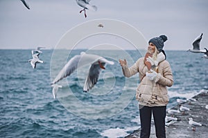 Happy young woman tourist feeds seagulls on the sea. Pretty female wearing coat, scarf, hat watching flying seagulls by the sea on