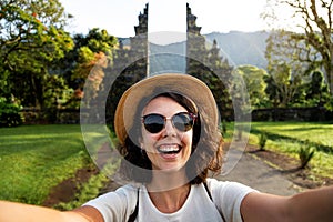 Happy young woman traveler looking at camera taking selfie in Balinese Hindu Temple entrance during summer vacation.
