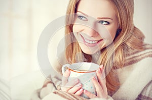 Happy young woman is under a blanket and cup of coffee on winter morning at home