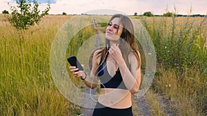 Happy young woman use smartphone and headphone standing in field at sunset, healthy lifestyle. Sportywoman runner using