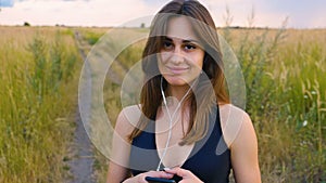 Happy young woman use smartphone and headphone standing in field at sunset, healthy lifestyle. Sportywoman runner using