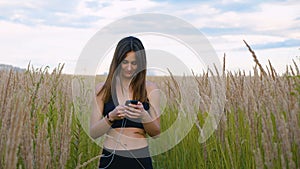 Happy young woman use smartphone and headphone walking in field at sunset, healthy lifestyle. Sportywoman runner using