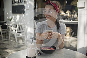 Happy young woman using her phone, having breakfast in cafe,smiles to somebody