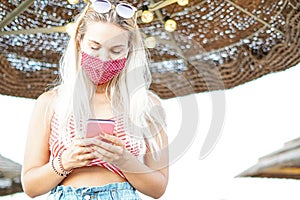 Happy young woman using mobile phone with face mask in beach - Teenager watching video in internet