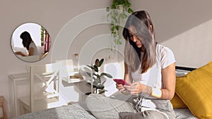 Happy young woman using smartphone sitting on bed in the morning at home.