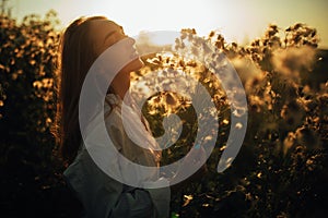 Happy young woman is walking among the flowers and grass on meadow at sunset