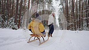 happy young woman is walking with her little child in winter forest, son is sitting in wooden sleigh