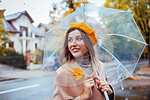 Happy young woman walking outdoors under transparent umbrella during rain holding yellow leaves. Fall season