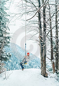 Šťastná mladá žena v teplom oblečení a červenej čiapočke hojdajúca sa na lesnej hojdačke s malebným výhľadom na zasnežené hory. Zimný čas
