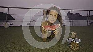 Happy young woman wearing bikini eating watermelon on the rooftop with beatiful view behind