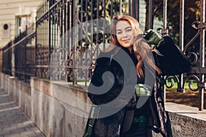 Happy young woman wearing fur coat with green handbag and gloves outdoors. Autumn clothes accessories. Female fashion