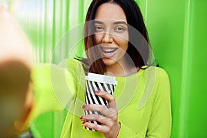 Happy young woman wearing green long sleeves shirt holding on the go, and taking self portrait on smartphone. Smiling girl