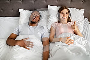 Happy young woman wearing headphones and showing thumb up gesture while her husband snoring on bed, top view