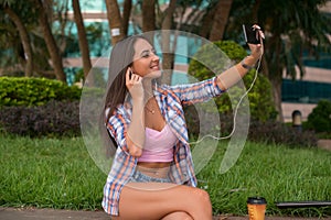 Happy young woman wearing headphones and taking a photo in the park sitting on the curb. Attractive female selfie.