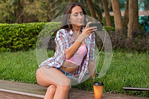 Happy young woman wearing headphones and taking a photo in the park sitting on the curb. Attractive female selfie.