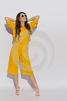 Happy young woman wearing a linen shirt, shorts and high heels poses with hands behind head