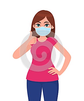 Happy young woman wearing medical face mask and showing thumbs up sign. Trendy girl covering protective surgical mask photo