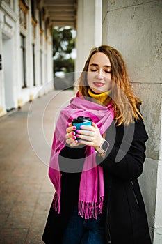 Happy young woman is wearing a pink scarf, She is holding a coffee to go. Portrait of smiling girl in autumn city.