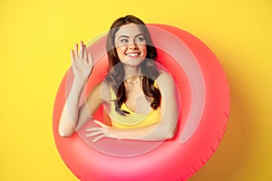 Happy young woman wearing pink swimming ring, waving hand and saying hi, enjoying vacation, summer beach holiday