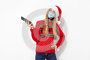 Happy young woman, wearing red santa claus hat and medical protective mask on face, with mobile phone, isolated on white