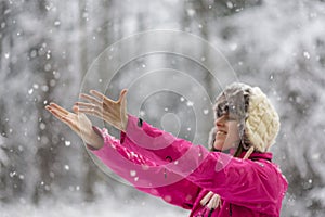 Happy young woman wearing warm hat and bright pink jacket standing outside in snowy nature