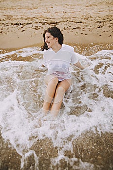 Happy young woman in wet white shirt lying on beach in splashing waves.  Emotional tanned girl relaxing on seashore and enjoying