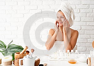 Happy young woman in white bath towels applying face cream doing spa procedures