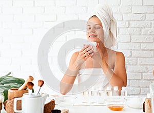 Happy young woman in white bath towels smelling the cream doing spa procedures