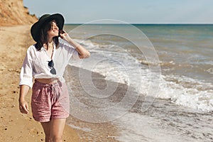 Happy young woman in white shirt and hat walking on sunny beach. Hipster slim girl relaxing near sea with waves, sunny warm