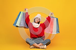 Happy young woman in winter clothes holding colorful shopping bags, sitting on yellow background