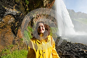 Happy young woman in yellow jacket posing under Seljalandfoss waterfall