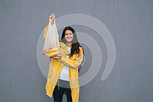 Happy young woman in a yellow raincoat holding string bag with fresh bananas and apples on the street against gray wall