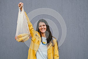 Happy young woman in a yellow raincoat holding string bag with fresh bananas and apples on the street against gray wall