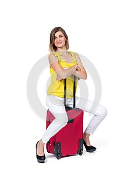 A happy young woman in a yellow T-shirt and white trousers is sitting on a red suitcase waiting, isolated on a white background