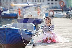 A girl in a striped sailor`s t-shirt and pink skirt sits alone on a wooden pier near the sea and boats