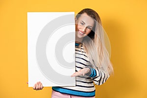 Happy young women holding white board, isolated on yellow background. Gorgeous girl showing blank empty paper billboard with blank