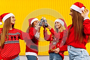 Happy young women in santa claus hats, friends cheerfully celebrate new year and drink drinks in glass bottles, on the background