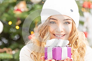 Happy youth blonde girl holds a well-packed present with a Christmas tree behind her