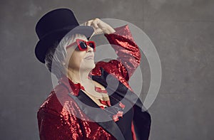 Happy youthful looking senior woman in top hat, cool glasses and sequin jacket posing in studio