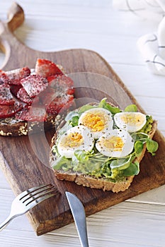 Hard boiled egg on avocado toast with green leaves and toast with fresh strawberries and cream cheese on wooden board, healthy bre