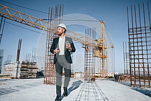 In hard hat, holding plan in hands. Businessman in formal clothes is on the construction site at daytime