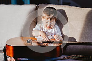 Hard light portrait of cute little baby boy playing big guitar sitting on sofa.