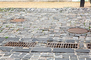 Hardscape sett road with iron grate of the drainage system hatch.