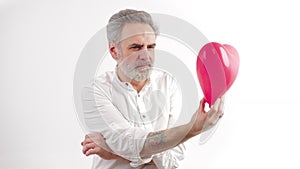 Hardships in love. Contemplating European bearded gray-haired middle-aged man in studio holding red heart balloon and