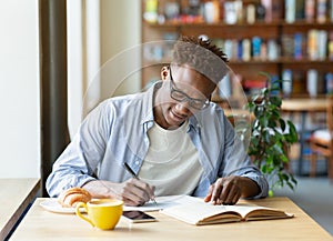 Hardworking black man writing down info from textbook, getting ready for test at cafe