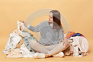 Hardworking Caucasian woman posing near heap of multicolored unsorted clothes isolated over beige background sorting her