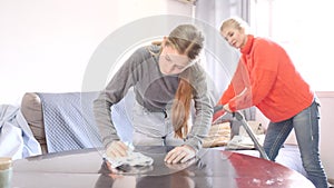 Hardworking daughter cleans the table with a rag while mother vacuums the room