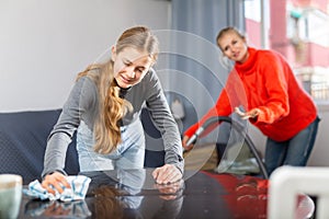 Hardworking daughter cleans the table with a rag while mother vacuums room