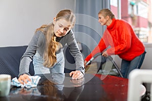 Hardworking daughter cleans the table with a rag while mother vacuums room